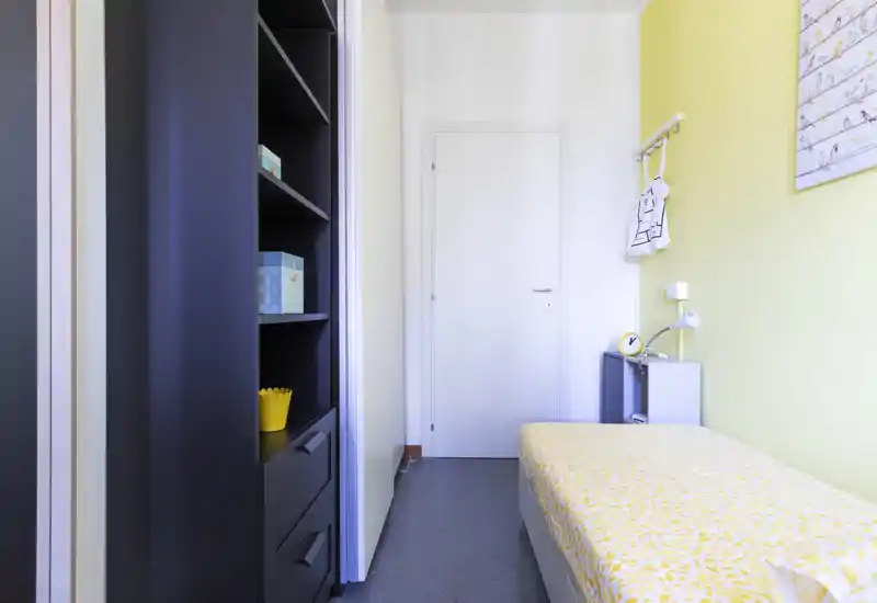 Piccola camera singola con copriletto giallo a motivo, mensola come comodino e appendiabiti a parete; luminosa, ordinata e arredata in modo essenziale, ideale per una persona.