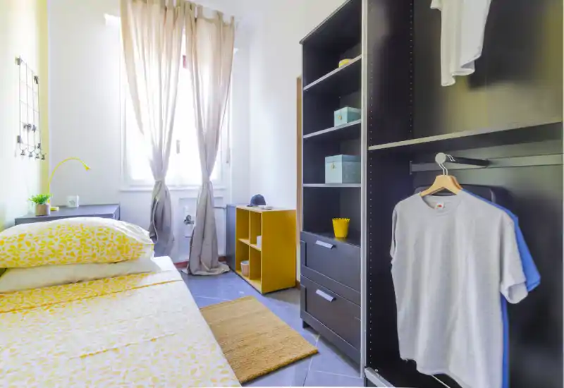 Camera da letto luminosa e compatta con biancheria gialla a motivi, luce naturale dalla finestra centrale e un armadio aperto. La stanza è ordinata e ben arredata con accessori coordinati e soluzioni di deposito pratiche.