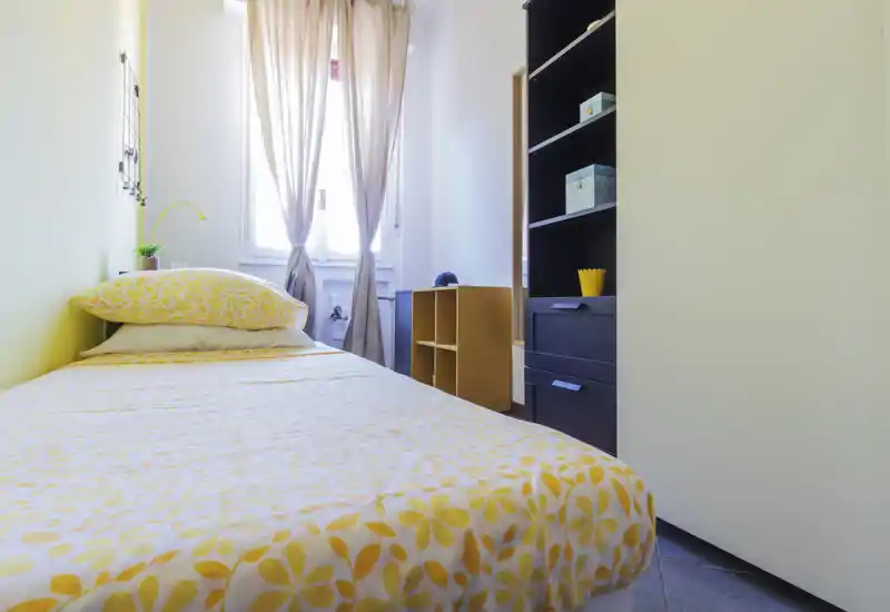 Camera da letto singola luminosa e ordinata con copriletto giallo a fiori, lampada e scaffalatura; luce naturale dalla finestra che rende l'ambiente fresco e accogliente.