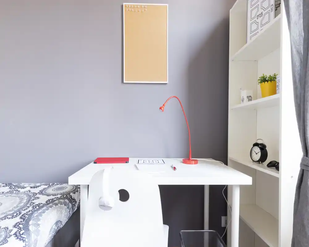 Compact bedroom corner with a white study desk, red desk lamp and a bed partially visible at left; shelving and a window with curtain on the right provide storage and natural light.