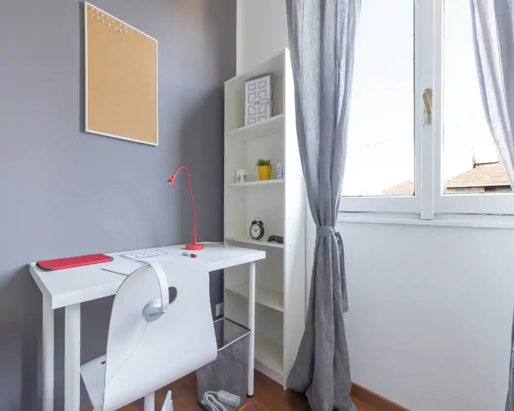 Bright, minimal home office corner with a white desk, task lamp, shelving and a large window letting in natural light—simple and functional workspace.