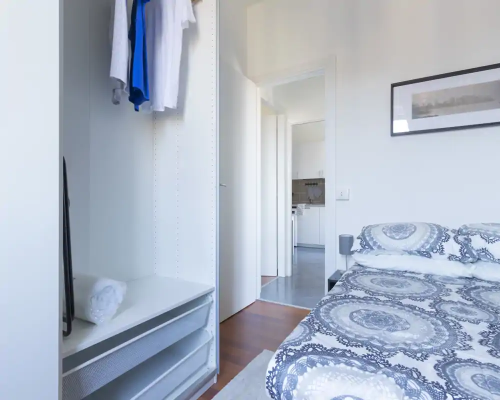 Bright, modern bedroom with patterned bedding, a bedside table and an open wardrobe; neutral tones and neat styling create a calm, airy atmosphere.