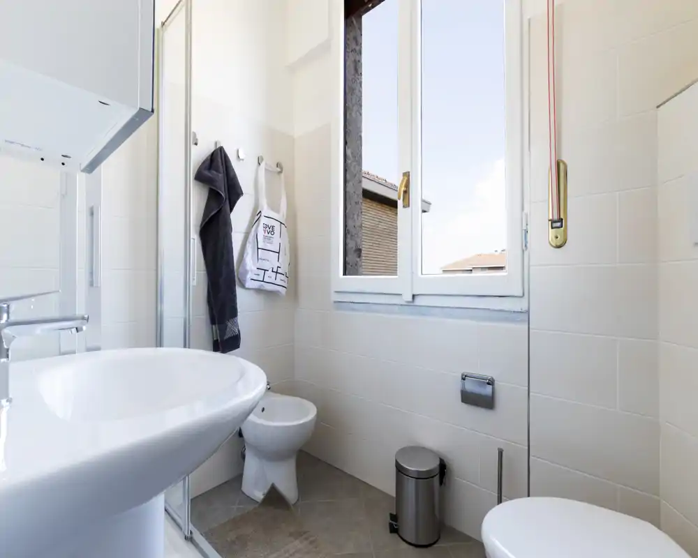 Bagno moderno e luminoso con lavabo a colonna, bidet e WC accanto a una finestra grande che illumina l’ambiente; piastrelle neutre e accessori ordinati creano uno spazio fresco e funzionale.