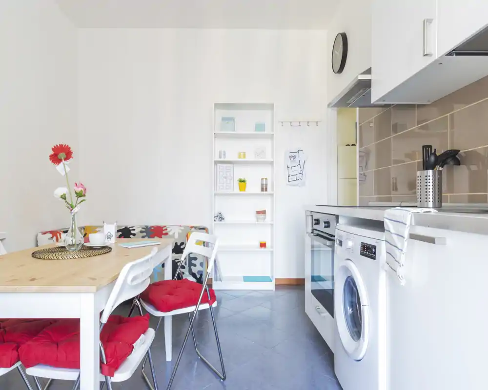 Cucina moderna compatta con elettrodomestici integrati, tavolo da pranzo con cuscini rossi e fiori decorativi che creano un ambiente luminoso e accogliente.