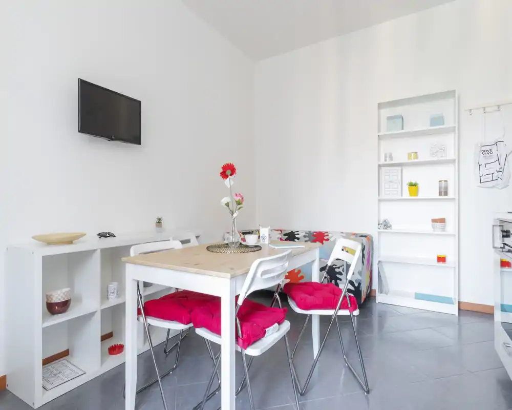 Angolo living/pranzo luminoso e moderno con un tavolo in legno, cuscini colorati e un divano compatto; scaffalatura minimal e TV a parete creano un ambiente pulito e accogliente.
