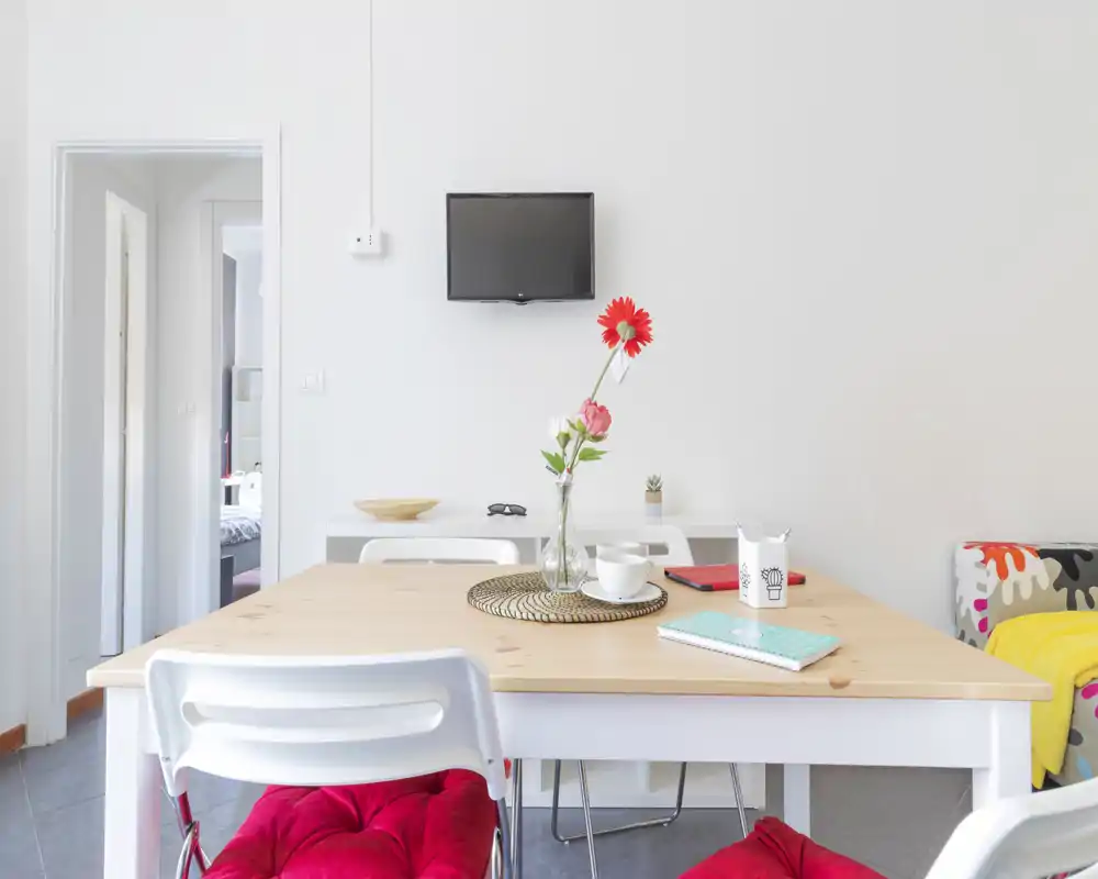 Zona pranzo moderna e luminosa con tavolo in legno, vaso con fiori, cuscini colorati e una piccola TV a parete — perfetta per annunci di appartamenti compatti.