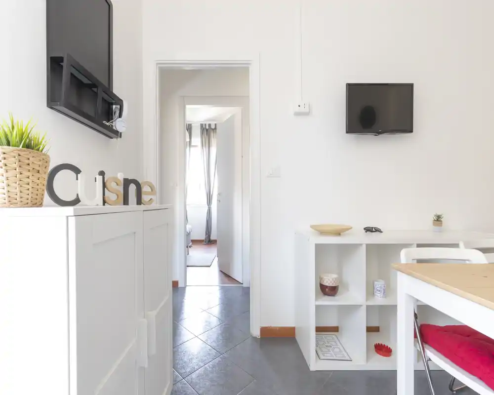 Zona pranzo luminosa e compatta con piccolo tavolo e scaffalatura, arredi semplici in stile nordico e luce naturale dalla stanza adiacente.