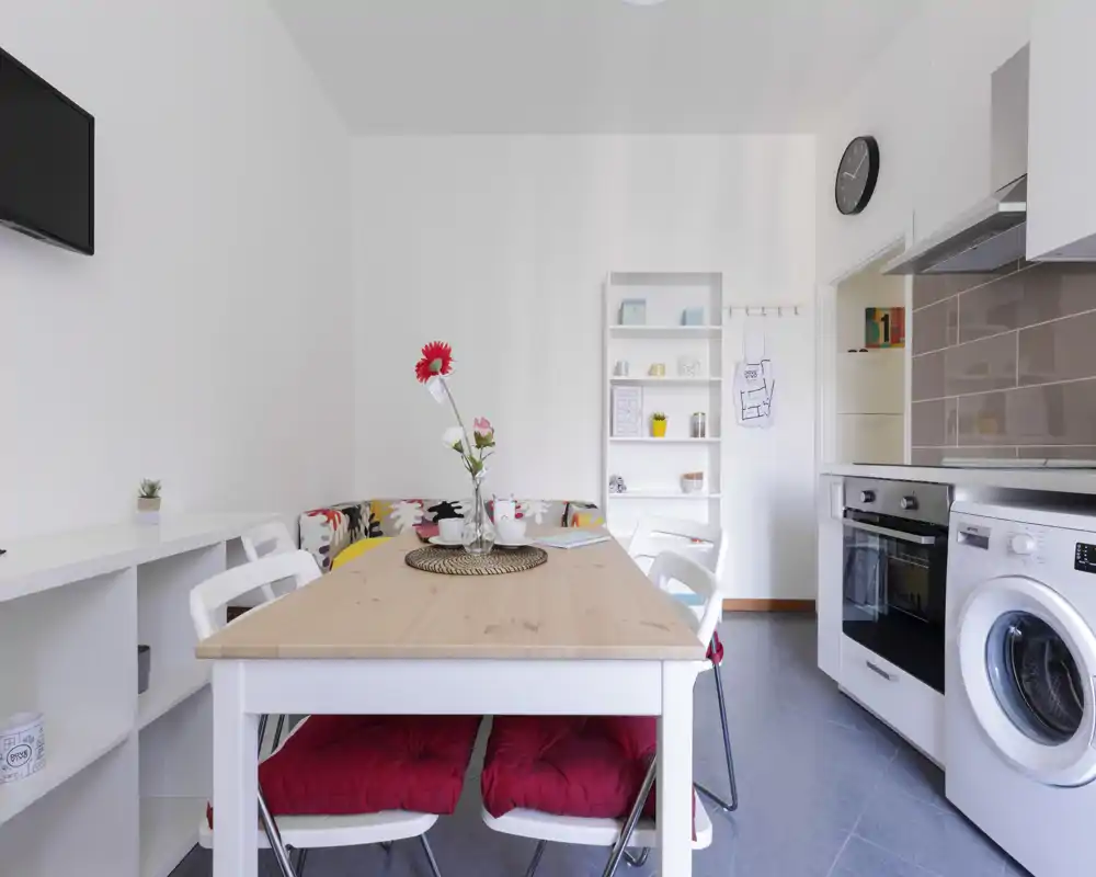Cucina moderna e luminosa con zona pranzo integrata: tavolo in legno con cuscini, elettrodomestici compatti (forno e lavatrice) e scaffalature ordinate che ottimizzano lo spazio.