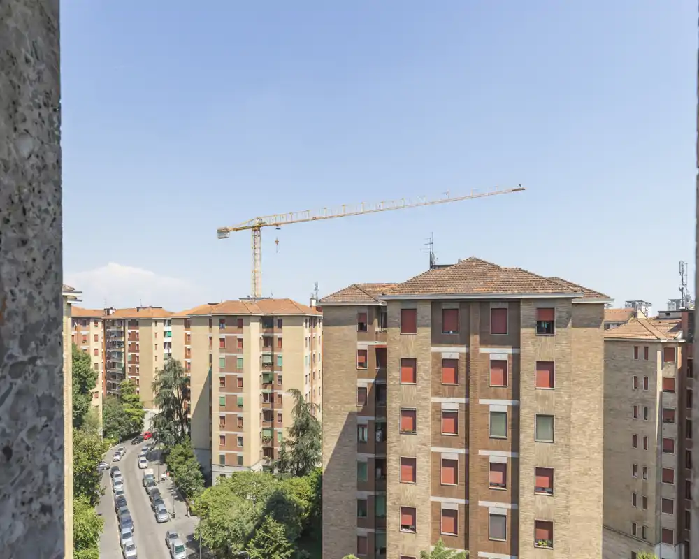 Vista cittadina da un piano alto con edifici residenziali in mattoni, una strada con auto parcheggiate, alberi e una gru da costruzione sotto un cielo limpido.