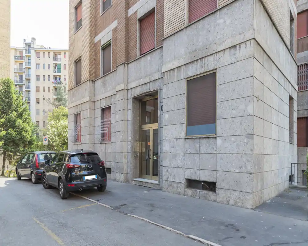 Vista esterna dell'ingresso di un palazzo residenziale e del parcheggio su strada adiacente, con marciapiede, auto parcheggiate e dettagli della facciata alla luce naturale.