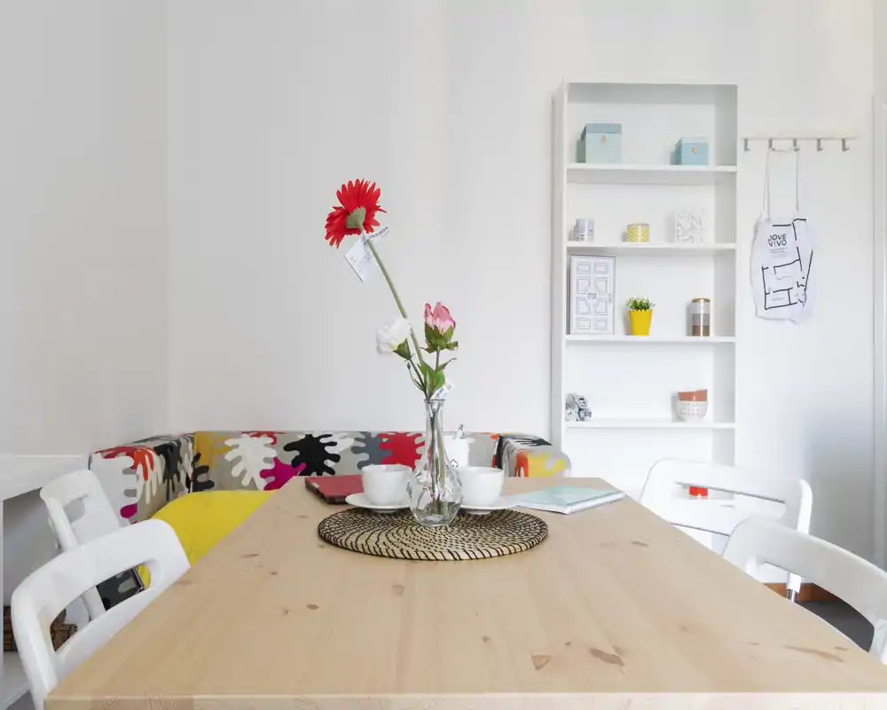 Angolo pranzo luminoso e moderno con tavolo in legno apparecchiato per due, vaso decorativo con fiori e panca imbottita colorata che rende lo spazio accogliente.