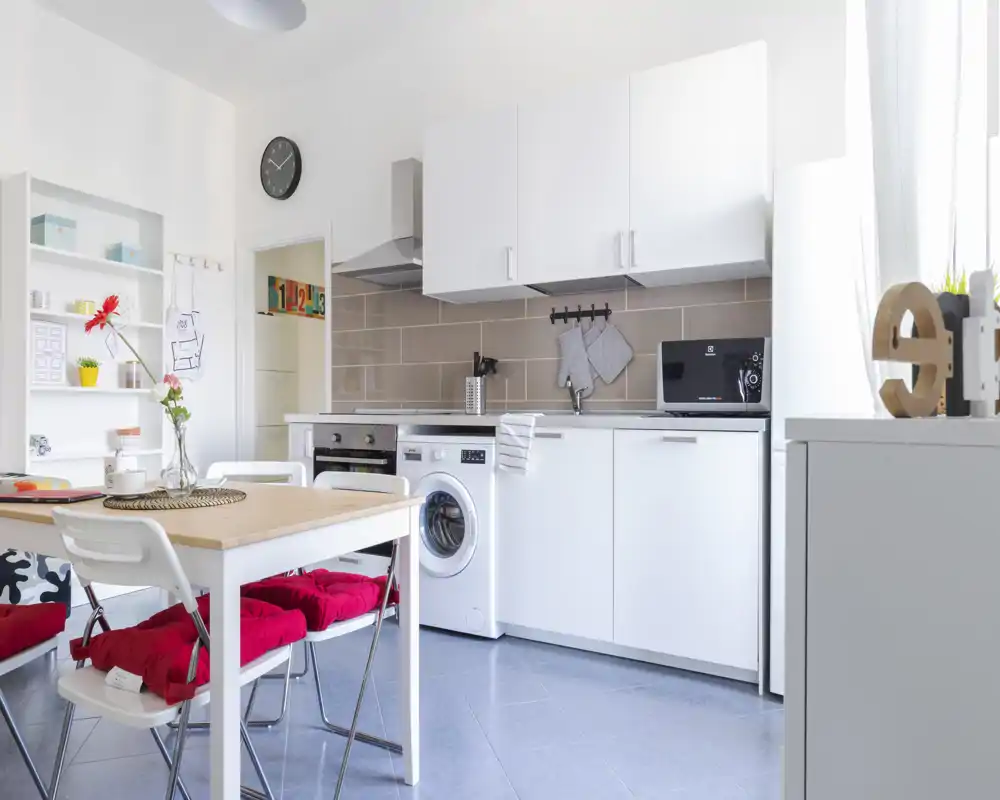 Cucina moderna luminosa con mobili bianchi, elettrodomestici integrati e un piccolo tavolo da pranzo con cuscini rossi; linee pulite e layout pratico, perfetto per un appartamento compatto.