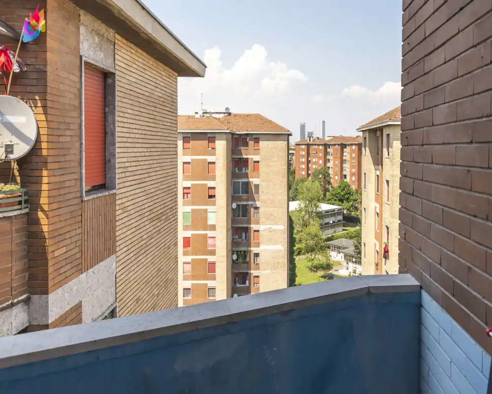 Piccolo balcone soleggiato con parapetto dipinto di blu con vista su condomini in mattoni e un cortile alberato.