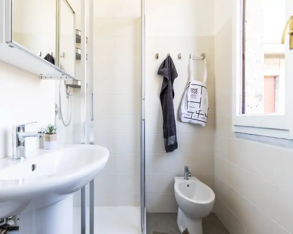 Bagno moderno e luminoso con lavabo su colonna, bidet, box doccia e una finestra che garantisce luce naturale. Piastrelle pulite e arredi minimali creano un ambiente funzionale e arioso.