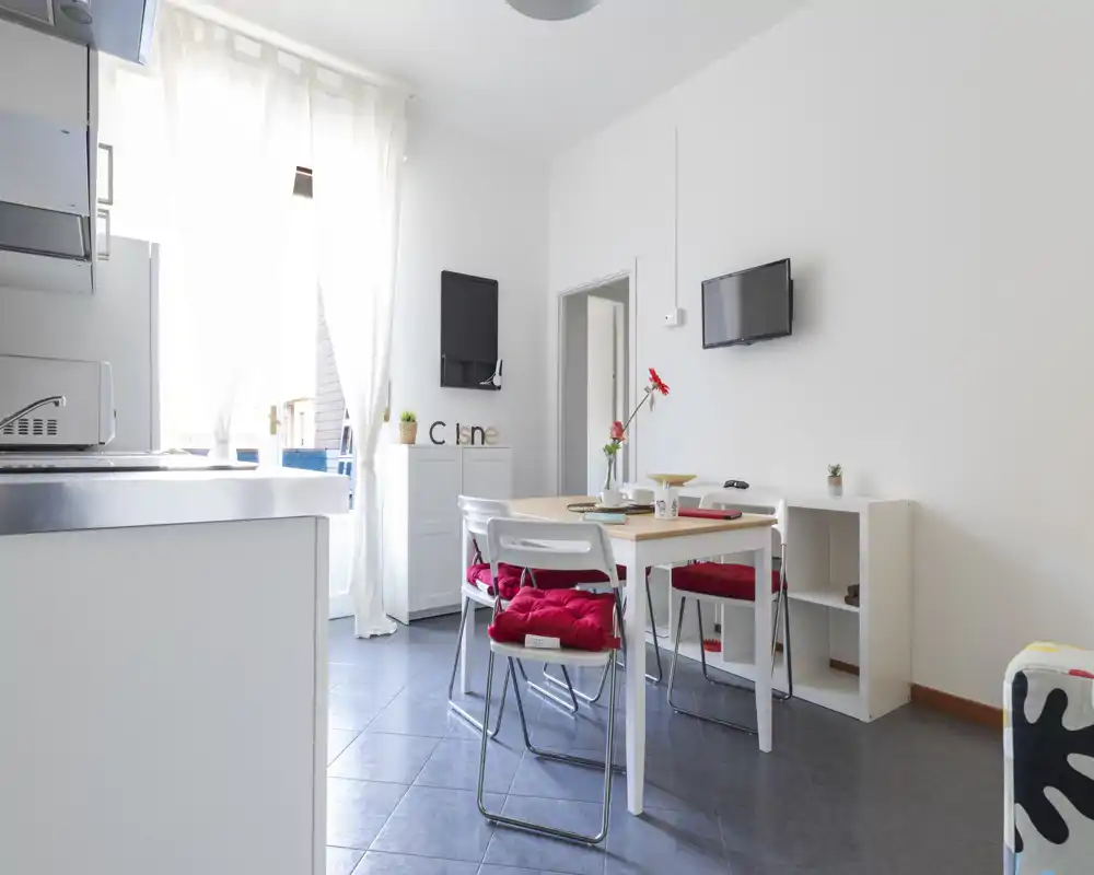 Zona pranzo luminosa e moderna con tavolo per quattro, cuscini rossi a contrasto e grande finestra che illumina l'ambiente.