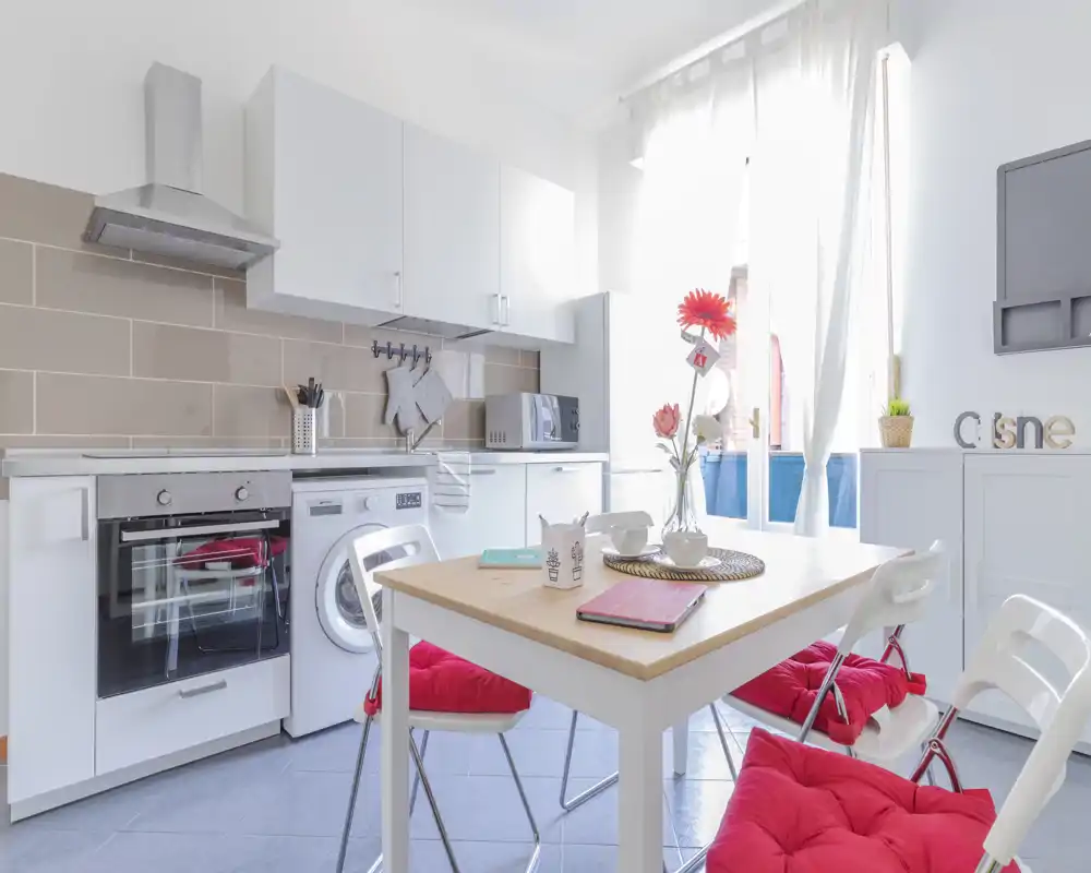 Cucina moderna e luminosa con ante bianche, forno incassato e lavatrice, tavolo da pranzo piccolo con sedie con cuscini rossi e decorazioni floreali.