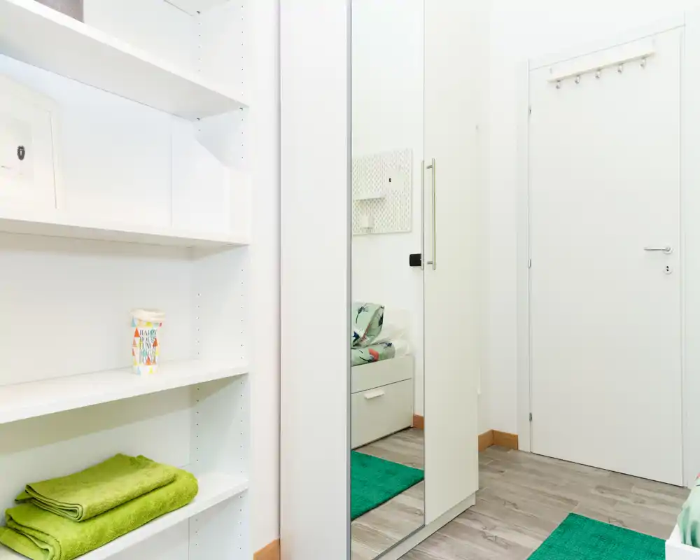 Compact, bright bedroom corner with white shelving and a mirrored wardrobe reflecting part of the bed; green textiles and rugs add a fresh accent.