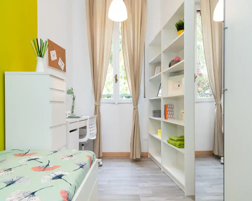 Bright, well-styled bedroom with a single bed, built-in shelving and a small study desk by the window. Neutral curtains and natural light create an airy, welcoming atmosphere ideal for a student or single occupant.
