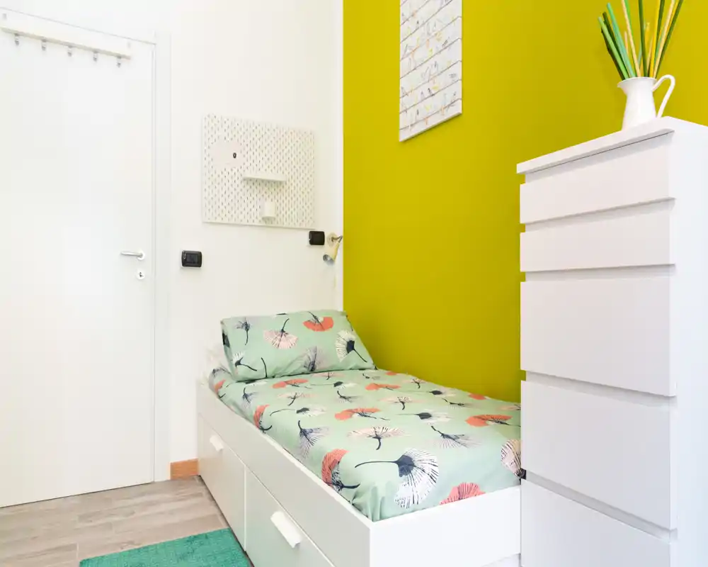 Compact single bedroom with a white storage bed, floral-patterned bedding and a tall white chest of drawers against a vivid green accent wall — bright, tidy and well styled for a rental listing.