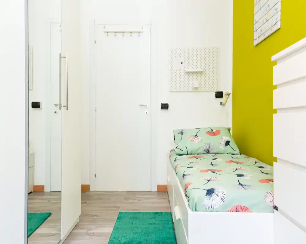 Small, bright bedroom with a single bed dressed in patterned bedding, a green accent wall and clean, modern storage. The room is well lit and styled for a cozy single occupancy.