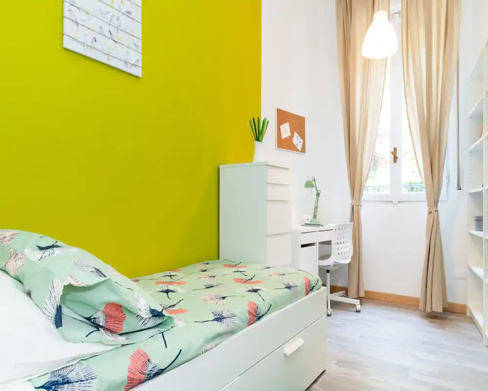 Bright, modern single bedroom with a patterned bed, compact white desk and chest of drawers beside a large window letting in natural light.