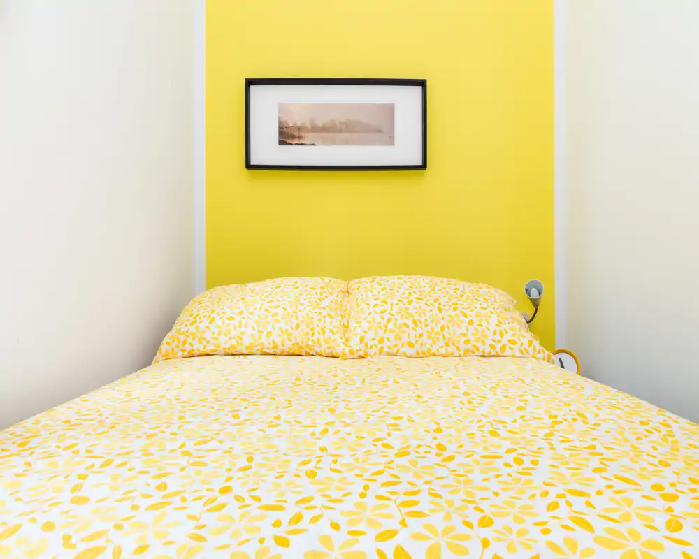 Chambre lumineuse et minimaliste avec un lit complet, linge de lit jaune à motifs sur une tête de lit jaune vif et un cadre au mur — propre, gaie et bien éclairée.