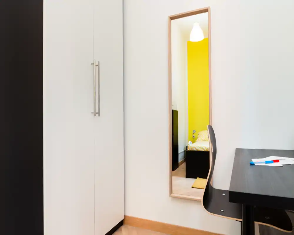 Détail d’une chambre photographié via un miroir : on aperçoit un lit compact avec une couette à motifs, tandis qu’une armoire moderne et un bureau occupent le premier plan.