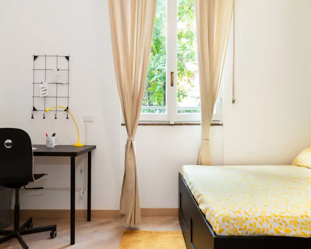 Chambre lumineuse et soignée avec un lit double partiellement visible habillé d'un linge de lit jaune à motifs, un petit bureau noir avec lampe jaune et une fenêtre laissant entrer la lumière naturelle.