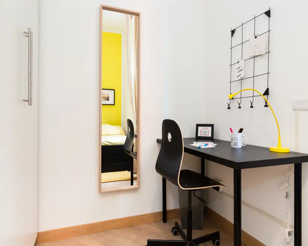 Coin de chambre lumineux vu dans un miroir intégral, avec un bureau et une chaise simples au premier plan ; un mur jaune et une literie chaleureuse apportent une touche cosy.
