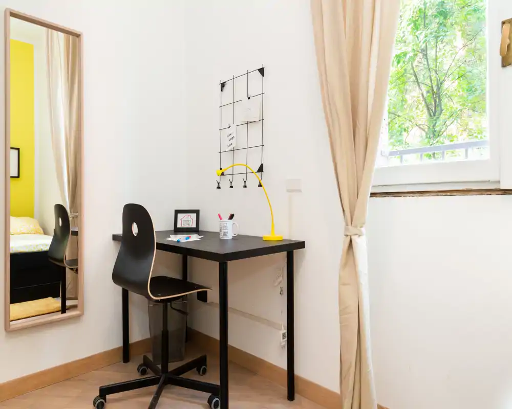 Coin chambre lumineux et soigné avec bureau, lampe jaune et miroir reflétant le lit — idéal pour une personne ou un étudiant.