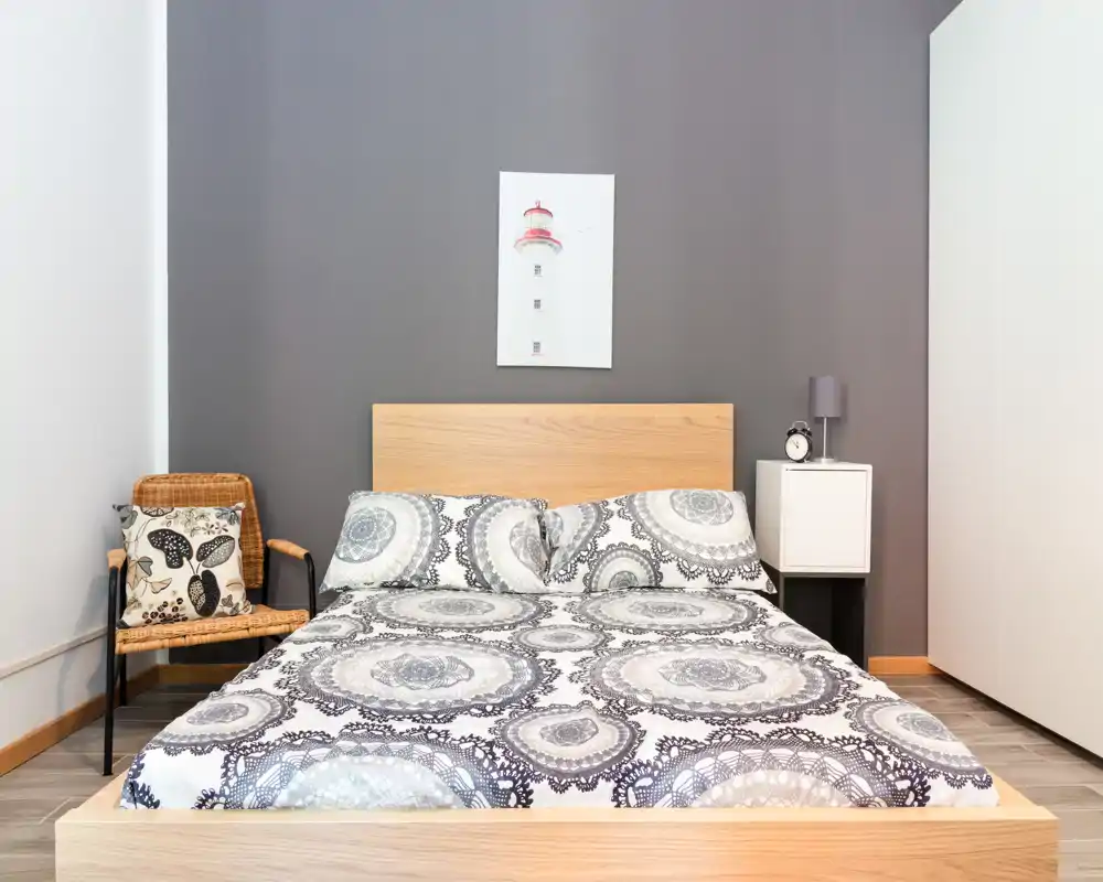 Dormitorio moderno y luminoso con cama grande de madera, ropa de cama estampada, mesita con lámpara y despertador y una silla de mimbre — limpio y de estilo sencillo para alquiler.