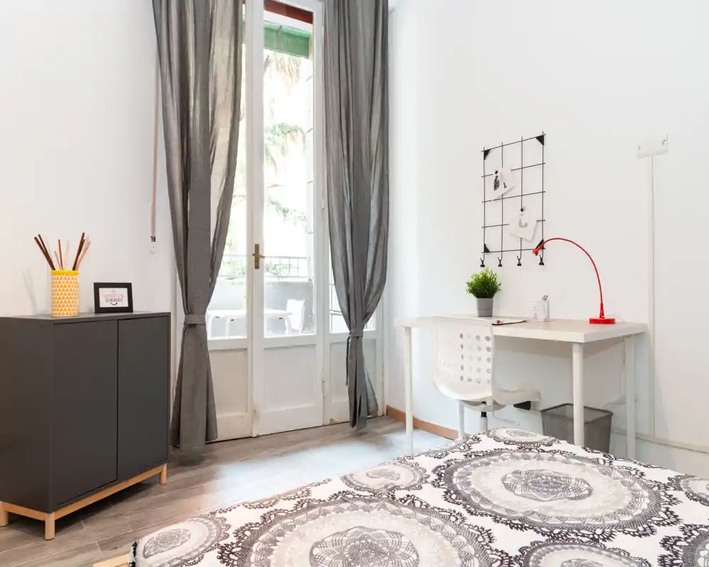 Dormitorio luminoso y minimalista con ropa de cama estampada, un escritorio pequeño y puertas al balcón que aportan luz natural. Líneas limpias y tonos neutros crean un espacio acogedor perfecto para alquileres temporales.