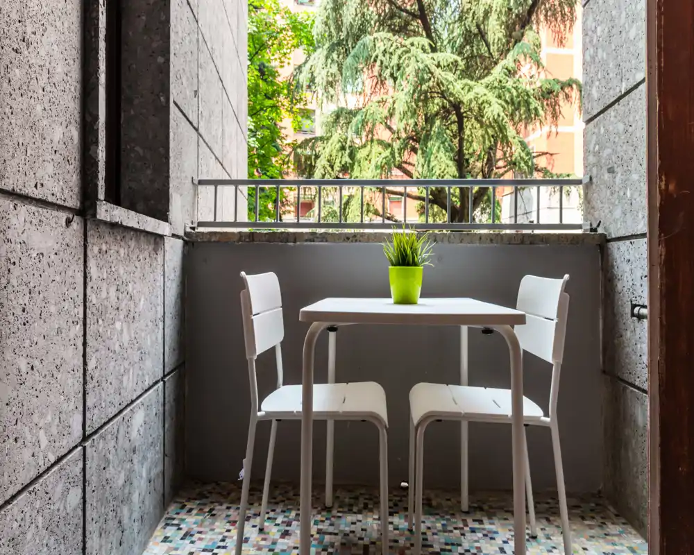 Pequeño balcón urbano con una mesa blanca compacta y dos sillas, una planta en maceta verde sobre la mesa y árboles de la ciudad visibles más allá de la barandilla.