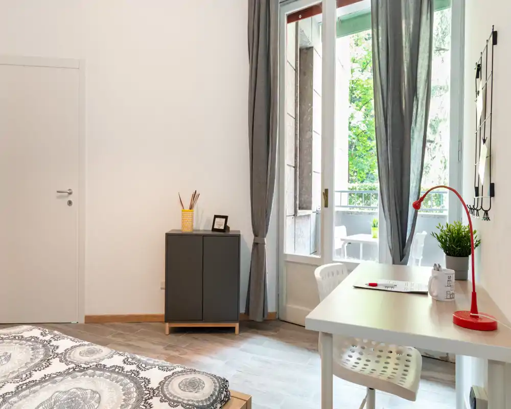 Dormitorio luminoso y ordenado con ropa de cama estampada, un escritorio junto a la ventana y una puerta al balcón que aporta luz natural.