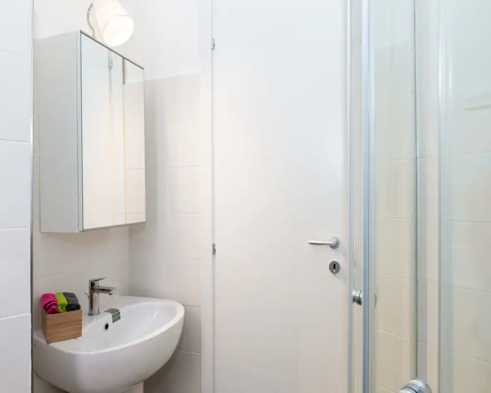 Clean, compact bathroom with a wall-mounted sink, mirrored medicine cabinet, and a glass shower door — bright white tiles and minimal, modern fixtures.