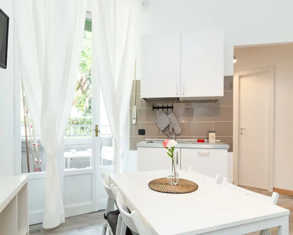 Bright, compact open-plan kitchen with a small dining table, white cabinets and a vase with fresh flowers creating a welcoming atmosphere.