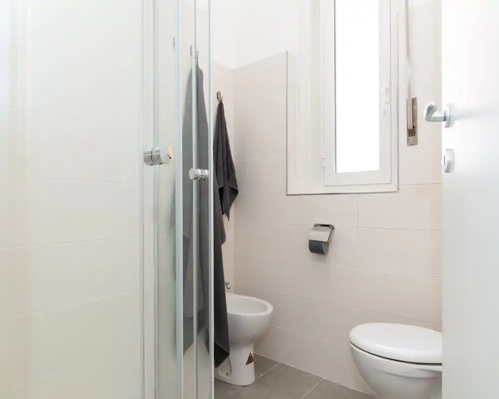 Clean, compact bathroom with a shower cabin, toilet and bidet. Neutral tiles and a window provide soft natural light.