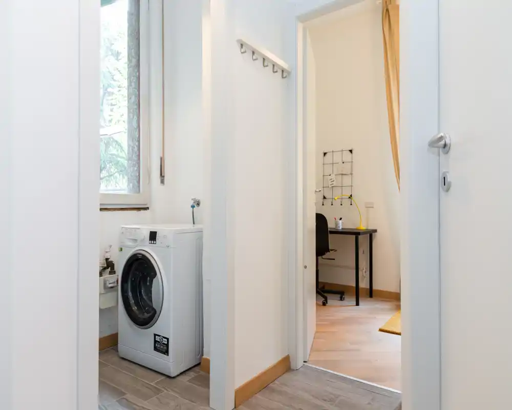 Small transitional entrance/hallway with a visible washing machine and a glimpse into an adjacent room with a desk; bright white walls and wood-tone baseboards create a clean, functional space.