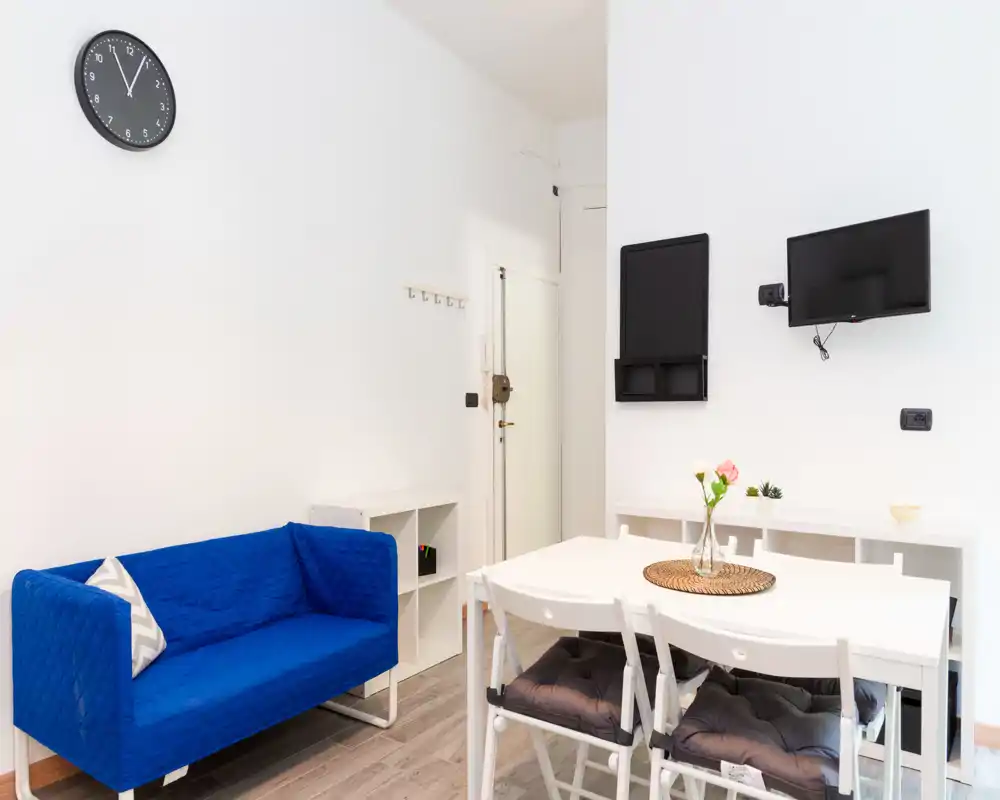 Bright, modern living/dining corner with a blue two-seater sofa, white dining table and wall-mounted TV — ideal for compact city stays.