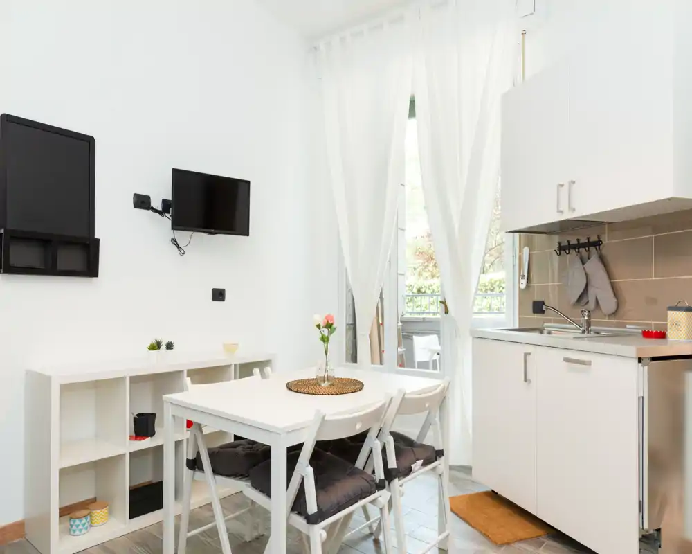 Bright, compact dining area with a white table and four chairs next to a small kitchenette and glass door leading to a balcony; simple, modern styling with decorative cushions and a fresh floral centerpiece.