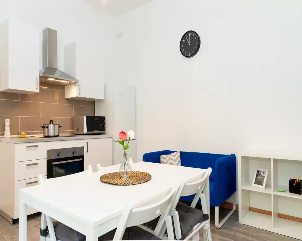 Bright, tidy open-plan living area with a compact kitchenette, white dining table and a blue two-seater sofa—ideal for a small apartment.