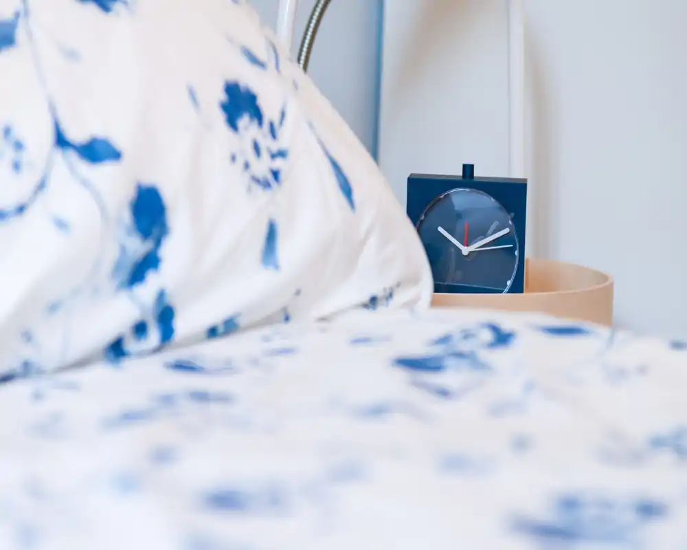 Primo piano dell'area letto con biancheria a fantasia e una piccola sveglia blu sul comodino; luce morbida e dettagli tessili accoglienti.