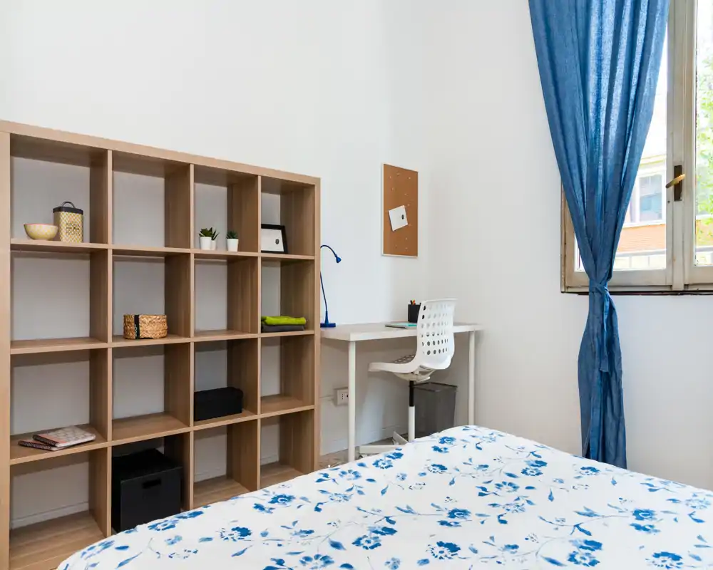 Camera da letto luminosa e minimal con copriletto floreale, scaffalatura a cubi e una scrivania vicino alla finestra — ambiente pulito e arioso, ideale per una singola persona o studente.