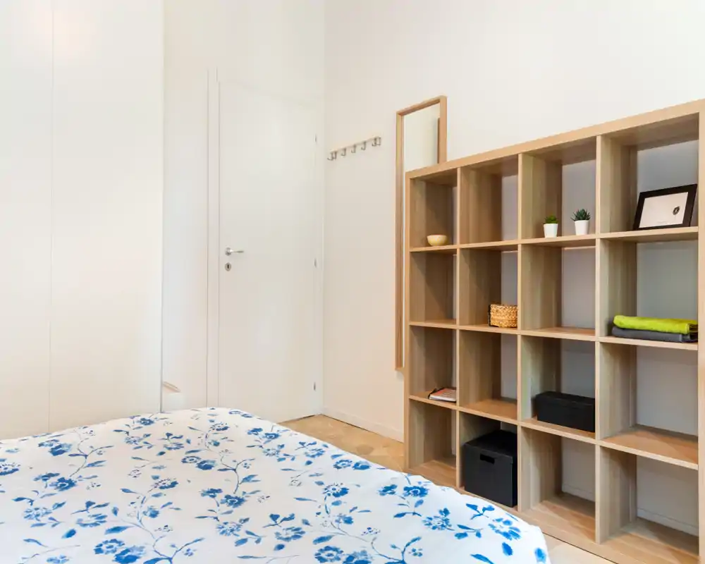 Camera da letto luminosa e minimal con copriletto a fiori e un mobile a cubi aperto; pareti neutre e pavimento in legno chiaro creano un’atmosfera ariosa e tranquilla.
