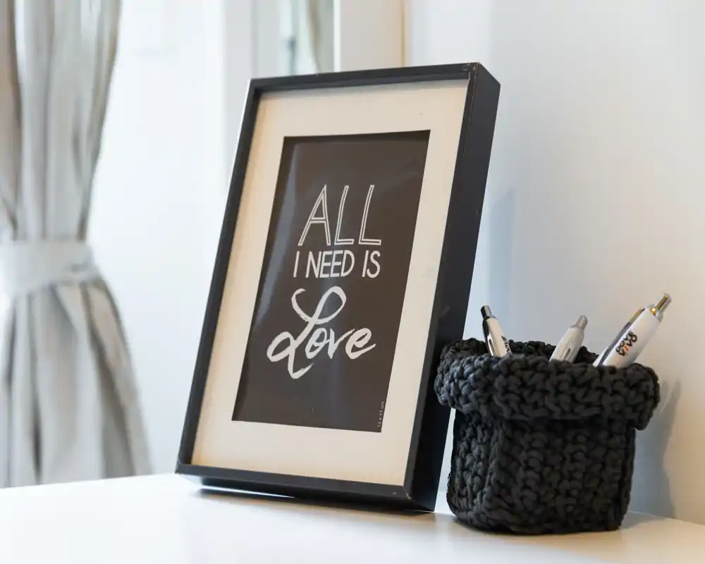 Primer plano de un marco con una lámina sobre una superficie blanca y un portabolígrafos tejido; al fondo se ve una cortina. La foto enfoca detalles decorativos, no la habitación completa.