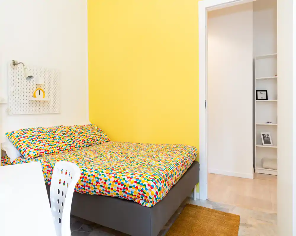 Dormitorio compacto y luminoso con una cama doble colorida frente a una pared amarilla; líneas limpias y decoración minimalista que crean un espacio acogedor, ideal para estancias cortas.