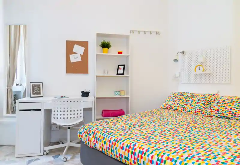 Dormitorio luminoso y moderno con una cama doble de estampado colorido, escritorio y estantería — ideal para un alquiler compacto y alegre.
