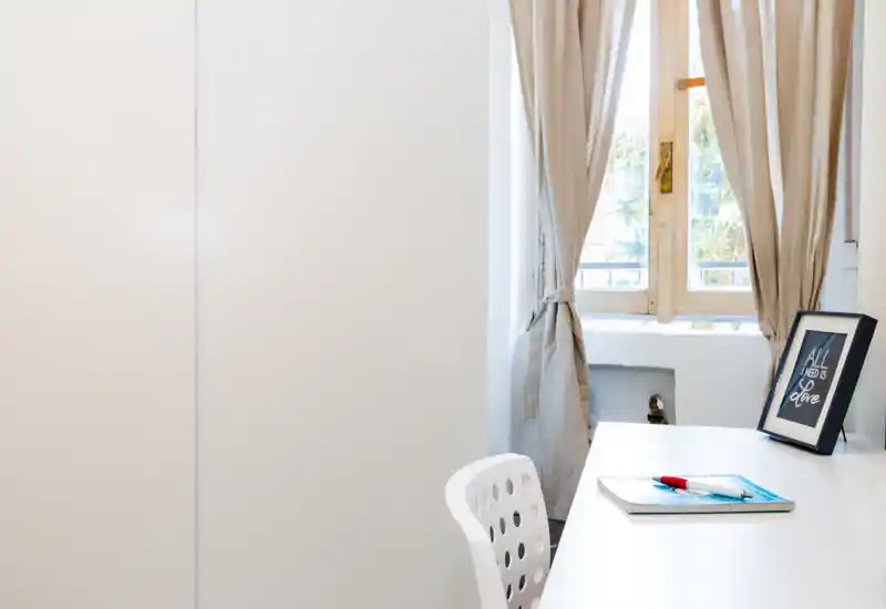 Rincón de sala luminoso y ordenado con escritorio blanco junto a la ventana, cortinas suaves y un marco decorativo; ideal como espacio de trabajo.