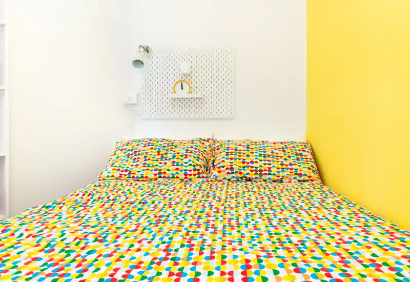 Dormitorio luminoso y colorido con una cama que domina la imagen y ropa de cama de estampado divertido; decoración mínima y lámpara de mesita.
