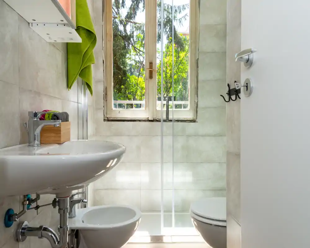 Baño pequeño y luminoso con lavabo colgante, bidé e inodoro, y mampara de ducha de cristal frente a una ventana que deja entrar la luz natural.
